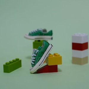 May include: A pair of miniature green canvas sneakers with white laces and soles. The sneakers are displayed on colorful toy building blocks against a light green background. One sneaker is leaning on a red and yellow block.