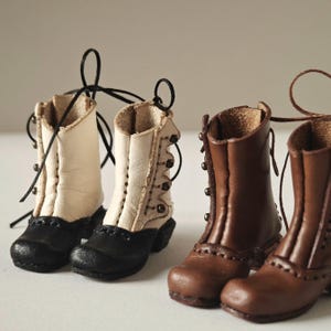 Puede incluir: Botas de cuero en miniatura en dos estilos. Un par es de color crema con punteras negras, el otro marrón. Ambos pares tienen cordones y tacones pequeños. Las botas son probablemente para una muñeca u otra figura pequeña.