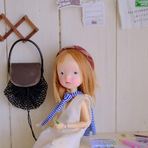 Puede incluir: Una muñeca con cabello rojo, que lleva una diadema a cuadros rojos y marrones, una bufanda a rayas azules y blancas y un vestido blanco. Un bolso marrón cuelga de un perchero de madera. Una bolsa de red negra está unida al bolso.