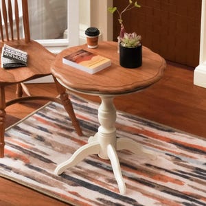 Może przedstawiać: Mały, okrągły stolik boczny z jasną podstawą i ciemnym drewnianym blatem. Na stole stoi mała czarna filiżanka do kawy, książka i doniczkowy sukulent. Widoczne są również drewniane krzesło i wzorzysty dywan.