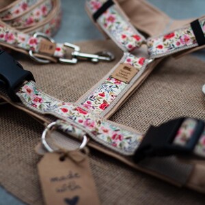 Puede incluir: Un arnés para perros y un juego de correa con diseño floral. El arnés tiene una base beige y está decorado con flores rojas, rosas y azules. Incluye una hebilla negra y un accesorio metálico para la correa. La etiqueta dice "made with love".