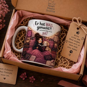 Könnte beinhalten: Eine weiße Keramiktasse mit einer Comic-Illustration einer Person, die mit einem Hund im Bett liest, und dem deutschen Text "Er hat WAS gemacht?! ...und warum finde ich das gut?". Enthält ein braunes Geschenketikett mit Text und eine Karte.