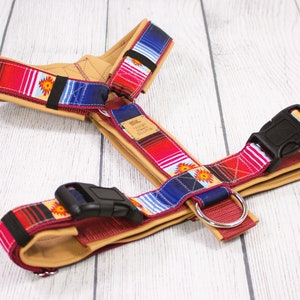 May include: A tan leather dog harness with red, blue, and white striped webbing. The webbing has a pattern of orange suns. The harness has two black plastic buckles and a red metal D-ring.