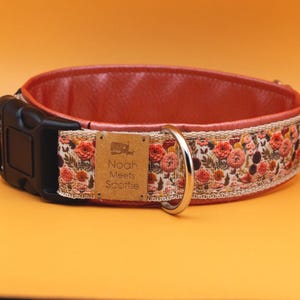 Op de afbeelding: Een koraalroze leren hondenhalsband met een bloemenprint stoffen rand en een zwarte gesp. De halsband heeft een gouden metalen D-ring en een gouden metalen label met de tekst "Noah Meets Scottie".