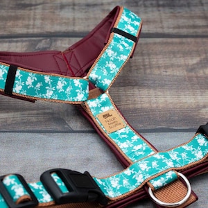 Può includere: Un'imbracatura per cani floreale blu-verde e bianca con un accento in pelle bordeaux e una fibbia nera. L'imbracatura ha un cinturino marrone con un anello di metallo e un'etichetta in pelle marrone che dice "Noah's Maine Scootie".