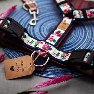 May include: A floral dog harness and lead set. The harness has a light green floral pattern with pink and red flowers, black buckles, and a brown interior. A tag reads "made with love."