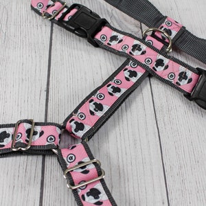 May include: A pink and grey dog harness with a black buckle and a silver ring. The harness has a pattern of small white and black dog faces on a pink background.
