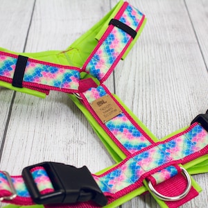 Puede incluir: Un arnés para perro verde lima con un patrón de escamas de sirena rosa y azul. El arnés tiene una hebilla negra y una etiqueta de cuero marrón que dice "Noon Meels Scootie".