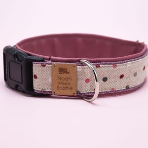 May include: A pink and beige dog collar with a white polka dot pattern. The collar has a black buckle and a silver D-ring. The collar has a brown leather patch with the text "Nooh Meets Scottie" on it.