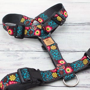 May include: A floral dog harness with a gray base and a colorful pattern of teal, yellow, and pink flowers. The harness has black plastic buckles and a metal ring for leash attachment. The brand name is visible on a small tag.