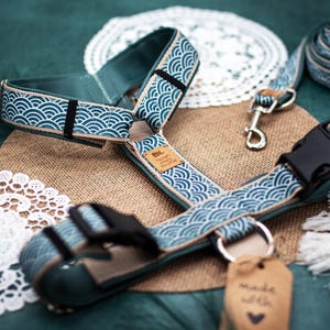 May include: A teal pet harness featuring a white wave pattern, black plastic buckles, and metal D-rings. A small tan label reads Noah Meets Scottie, and a brown paper tag attached to the harness displays the text made with a heart symbol.