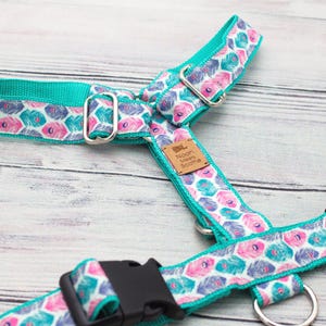 May include: A dog harness in teal with a pattern of pink, teal, and purple flowers. It features a black buckle and metal fixings. A small brown tag is attached to the harness.