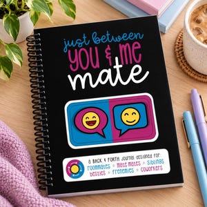 Puede incluir: Un diario de espiral negro con el texto "just between you & me mate" en azul, rosa y blanco. Presenta un gr&aacute;fico de dos burbujas de di&aacute;logo con caras sonrientes amarillas y una etiqueta que enumera "roommates, mate mates, siblings, besties, frenemies, coworkers."