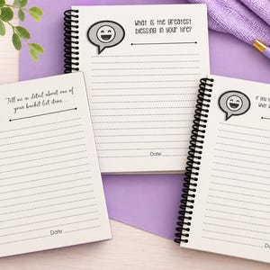 Puede incluir: Tres cuadernos de espiral con papel blanco rayado. Cada p&aacute;gina presenta un icono de burbuja de di&aacute;logo gris con una cara sonriente y una pregunta: "What is the greatest blessing in your life?", "Tell me in detail about one of your bucket list items", y "If you had to listen to one song forever, what would it be? Why do you love it?"