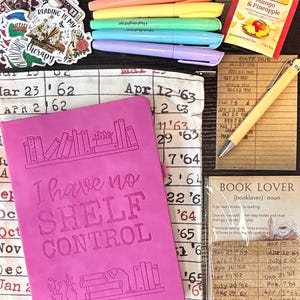 May include: A bright pink notebook embossed with the text I have no SHELF CONTROL and illustrations of books, a pencil case with a library due date card print, six pastel highlighters, a Twinings tea bag, a yellow pen, and several book-themed stickers.