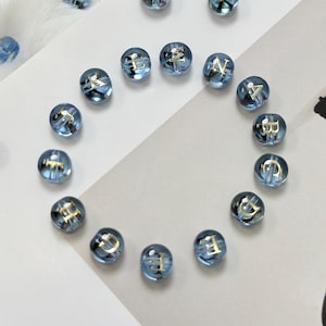 May include: A collection of clear blue glass beads with gold lettering. The beads are arranged in a circle and scattered across a white surface. Each bead has a different letter, suggesting they are for jewelry making or crafts.