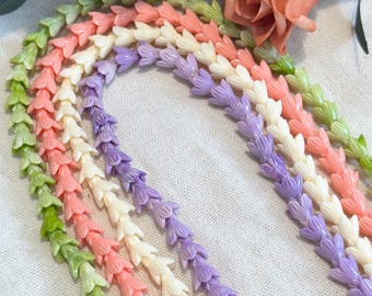 May include: A collection of five beaded necklaces, each featuring a strand of small, flower-shaped beads in pastel shades of light green, coral pink, cream, and lavender. The necklaces are arranged in a curved, overlapping pattern.