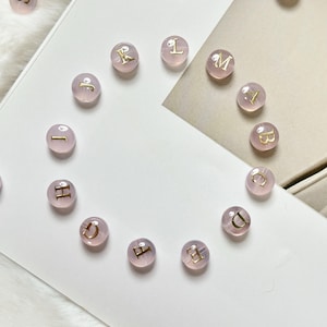 May include: Pink, translucent round beads with gold-colored letters from the alphabet. The beads are scattered on a white surface with a soft, fuzzy texture in the background. The letters are in a serif font.