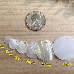 Op de afbeelding: Een verzameling witte, iriserende schelpen hangers van 10mm tot 30mm, liggen op een houten oppervlak. Een Amerikaanse munt wordt gebruikt ter vergelijking. De hangers zijn rond en hebben een gat om te rijgen.