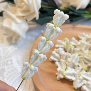May include: Close-up of white flower buds on a wire stem, with a wooden plate holding more flower buds. The buds have a delicate, layered petal design and a touch of yellow at the base. White roses are in the background.