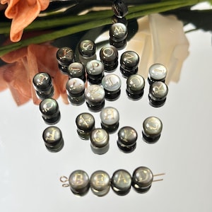 May include: A selection of dark, iridescent beads with gold lettering, spelling out letters of the alphabet. The beads are displayed on a reflective surface, with some strung together to spell "BEAD".