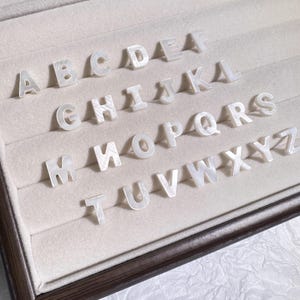 Peut inclure: Un ensemble de lettres de l'alphabet majuscules blanches et iridescentes, disposées dans un présentoir. Les lettres ont un aspect nacré et sont disposées en rangées. Le présentoir a un cadre en bois marron et un intérieur beige.