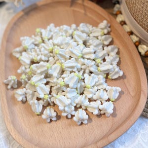 May include: A wooden tray filled with small, white, flower-shaped decorations with yellow centers. The decorations are clustered together, creating a textured surface. The tray is made of light-colored wood.