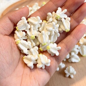 May include: A handful of small, white, flower-shaped craft embellishments with yellow tips. The delicate, three-dimensional floral pieces are clustered together, suggesting a craft supply or decorative element for DIY projects. The background is a wooden surface.