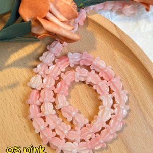 May include: A necklace of pink flower-shaped beads. The beads have a gradient effect, transitioning from a lighter to a darker pink. The necklace is displayed on a wooden tray, with artificial flowers in the background.