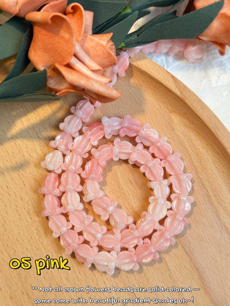 May include: A necklace of pink flower-shaped beads. The beads have a gradient effect, transitioning from a lighter to a darker pink. The necklace is displayed on a wooden tray, with artificial flowers in the background.