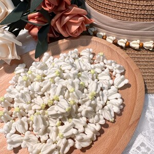 May include: A wooden tray filled with numerous white, flower-shaped decorations with yellow centers. Peach-colored roses and a straw hat with a beige ribbon are in the background. The decorations are likely for crafts or embellishments.