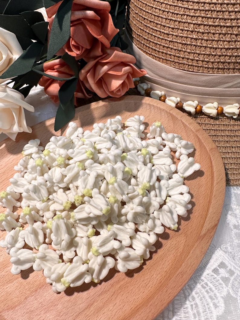 May include: A wooden tray filled with numerous white, flower-shaped decorations with yellow centers. Peach-colored roses and a straw hat with a beige ribbon are in the background. The decorations are likely for crafts or embellishments.