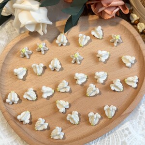 May include: A wooden tray holds numerous small, white flower-shaped embellishments with pale yellow centers. The flowers are arranged on a light brown wooden tray, surrounded by decorative flowers.