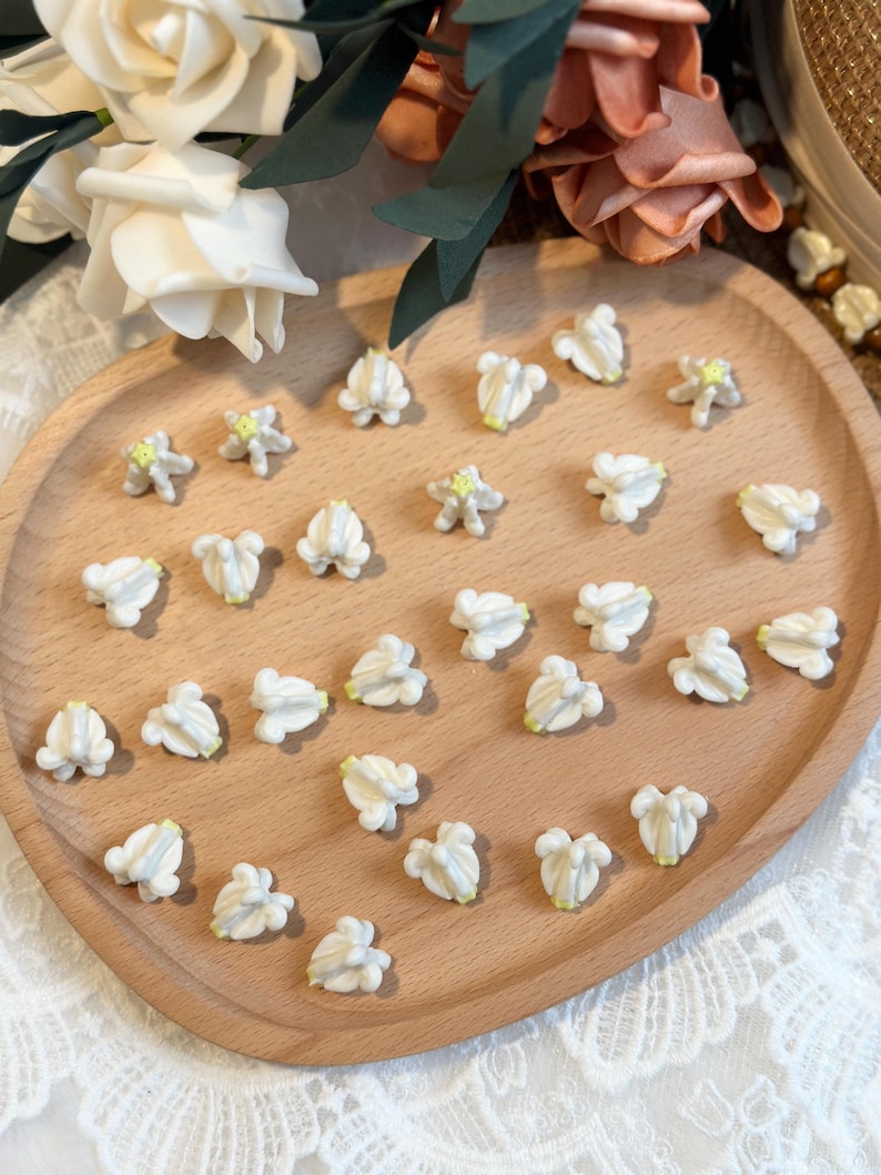 May include: A wooden tray holds numerous small, white flower-shaped embellishments with pale yellow centers. The flowers are arranged on a light brown wooden tray, surrounded by decorative flowers.