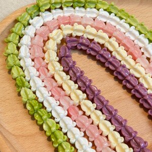May include: Rows of colorful frog-shaped beads in shades of green, white, pink, yellow, and purple. The beads are strung together, creating a textured pattern. The beads are displayed on a light brown wooden surface.