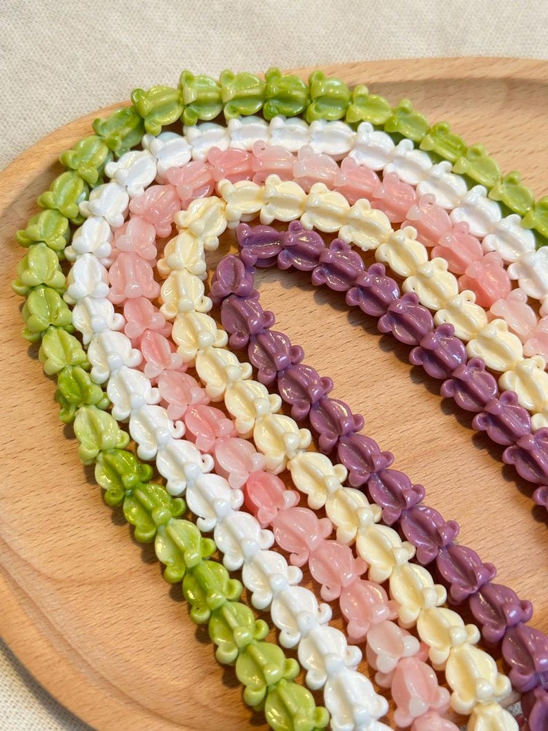 May include: Rows of colorful frog-shaped beads in shades of green, white, pink, yellow, and purple. The beads are strung together, creating a textured pattern. The beads are displayed on a light brown wooden surface.