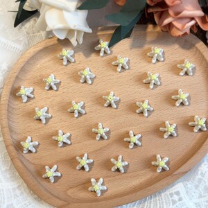 May include: A wooden tray holds numerous small, white, star-shaped embellishments with yellow centers. The tray is oval and made of light-colored wood. The background includes white and peach-colored flowers.