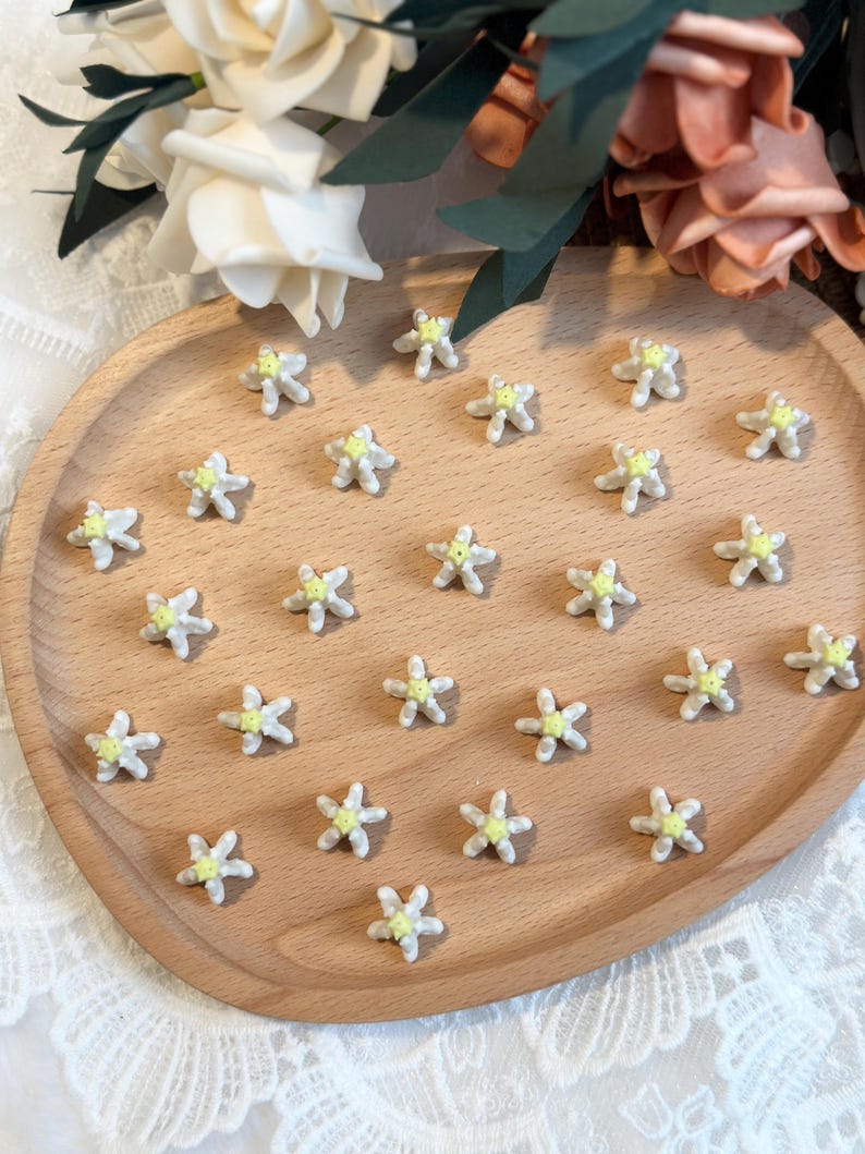 May include: A wooden tray holds numerous small, white, star-shaped embellishments with yellow centers. The tray is oval and made of light-colored wood. The background includes white and peach-colored flowers.