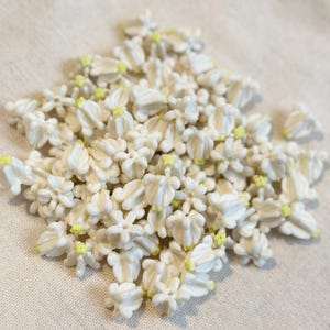 May include: A pile of small, white, flower-shaped decorations with yellow centers. The decorations are clustered together, creating a textured surface. The image is taken from a slightly elevated angle, providing a clear view of the details.