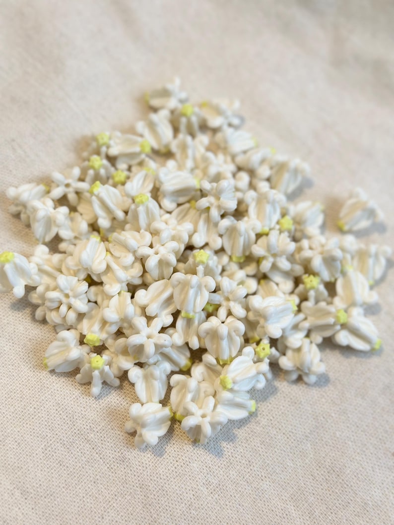 May include: A pile of small, white, flower-shaped decorations with yellow centers. The decorations are clustered together, creating a textured surface. The image is taken from a slightly elevated angle, providing a clear view of the details.