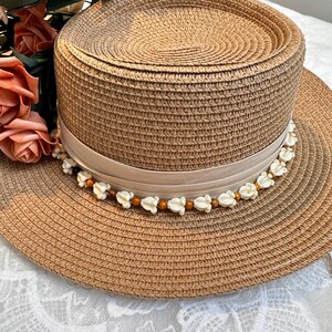 May include: A woven straw hat with a wide brim and a flat top. The hat features a beige ribbon around the crown, adorned with small white flower and brown bead accents. The hat is a light brown color and appears to be new.