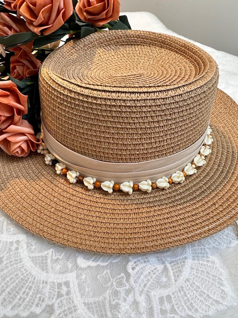May include: A woven straw hat with a wide brim and a flat top. The hat features a beige ribbon around the crown, adorned with small white flower and brown bead accents. The hat is a light brown color and appears to be new.