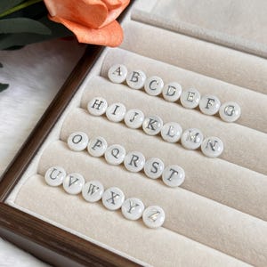 May include: White, round alphabet beads with silver lettering, arranged in rows inside a wooden jewelry box. The letters A through Z are displayed. A peach-colored flower is in the background.
