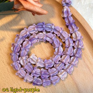 May include: A light purple necklace composed of flower-shaped beads. The beads are arranged in a circular pattern on a wooden tray, with a few beads extending upwards. The necklace is a soft, pastel shade of purple.