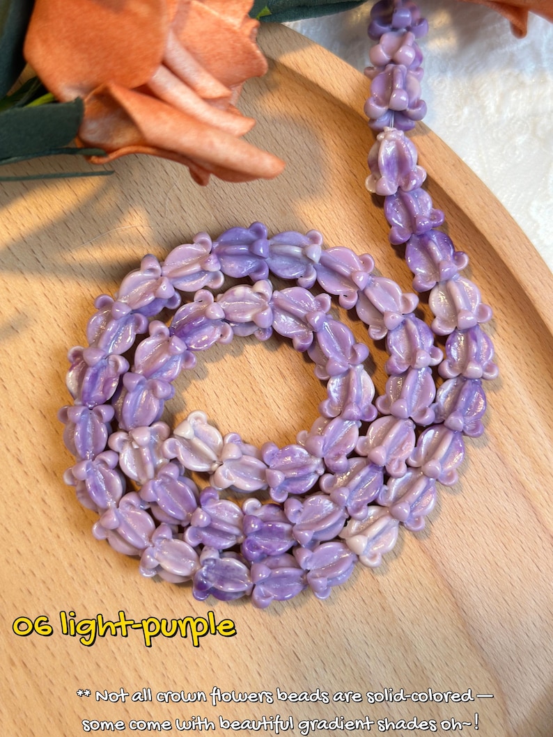May include: A light purple necklace composed of flower-shaped beads. The beads are arranged in a circular pattern on a wooden tray, with a few beads extending upwards. The necklace is a soft, pastel shade of purple.