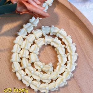 May include: Ivory-colored necklace composed of flower-shaped beads. The beads are arranged in a circular pattern on a wooden tray, with a peach-colored flower in the background. Text on the image reads "03 ivory".
