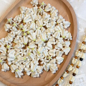 May include: A wooden tray filled with small, white, flower-shaped beads with yellow centers. A beaded necklace with white flower beads and brown wooden beads is visible on the right side. The beads are likely for crafting or jewelry making.