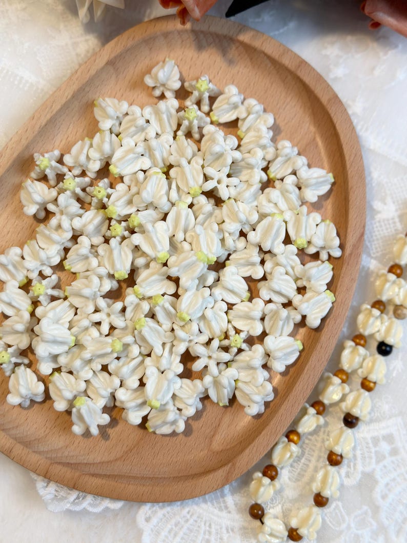May include: A wooden tray filled with small, white, flower-shaped beads with yellow centers. A beaded necklace with white flower beads and brown wooden beads is visible on the right side. The beads are likely for crafting or jewelry making.
