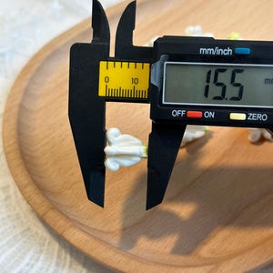 May include: A digital caliper measuring a small, white floral-shaped object. The caliper displays a measurement of 15.5 mm. The object is on a wooden tray, with a white lace fabric in the background.