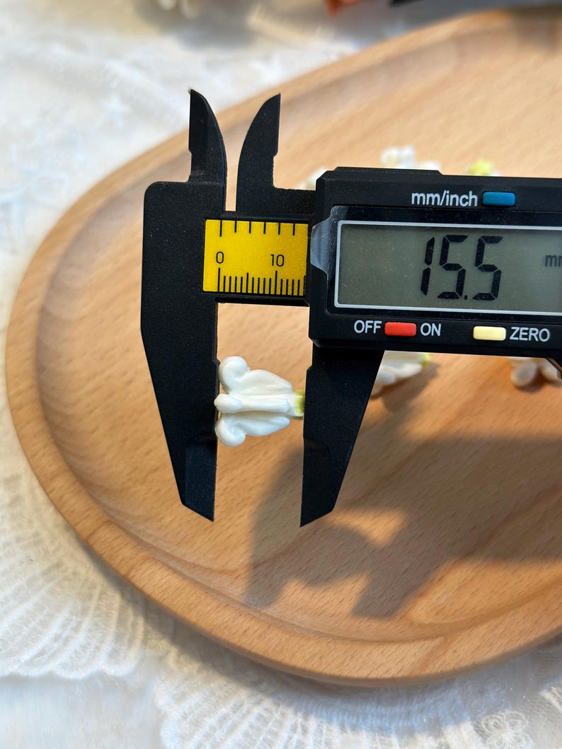 May include: A digital caliper measuring a small, white floral-shaped object. The caliper displays a measurement of 15.5 mm. The object is on a wooden tray, with a white lace fabric in the background.